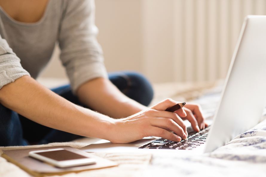 unrecognizable-young-woman-sitting-on-bed-working-on-laptop-smart-phone-next-to-her-ho-SBI-305226351